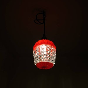 Puede incluir: Una l&aacute;mpara colgante de cristal rojo y transparente con un dise&ntilde;o facetado. La l&aacute;mpara tiene una parte superior e inferior rojas y cuelga de un cable negro. La luz est&aacute; encendida, proyectando un brillo c&aacute;lido en una habitaci&oacute;n oscura.