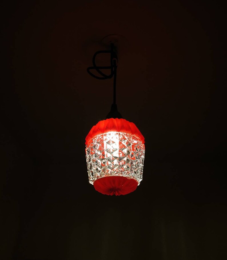 Puede incluir: Una l&aacute;mpara colgante de cristal rojo y transparente con un dise&ntilde;o facetado. La l&aacute;mpara tiene una parte superior e inferior rojas y cuelga de un cable negro. La luz est&aacute; encendida, proyectando un brillo c&aacute;lido en una habitaci&oacute;n oscura.