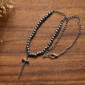 May include: A silver necklace with a sword pendant and a chain made of small, round beads. The necklace is lying on a wooden surface.