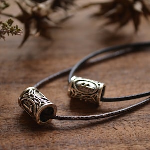 Op de afbeelding: Een ketting van bruin lederen koord met twee zilveren cilindrische kralen. Elke kraal heeft een ingewikkeld Keltisch knoopontwerp.