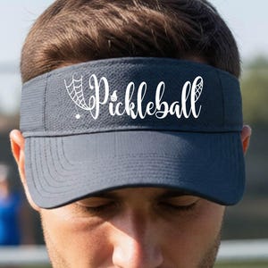 Könnte beinhalten: Ein dunkelblauer Sport-Visor mit dem weißen Schriftzug "Pickleball" in Schreibschrift, wobei eine kleine weiße Spinnennetz-Grafik in den ersten Buchstaben des Wortes integriert ist, platziert auf der Vorderseite des Visors.