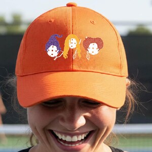 Könnte beinhalten: Eine leuchtend orangefarbene Baseballkappe mit einem Grafikdruck von drei stilisierten Figuren mit markanten Haarfarben in Lila, Gelb und Rot. Das Design ist mittig auf der Vorderseite der Kappe platziert, die einen gebogenen Schirm besitzt.