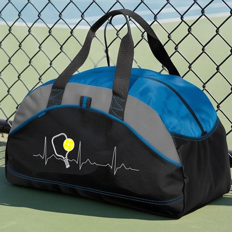 May include: A duffel bag featuring a blue, grey, and black colour scheme with two black handles. The front displays a white graphic of a heartbeat line integrated with a pickleball paddle and a yellow pickleball, centred on the black panel.