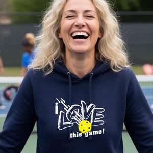 A navy blue hooded sweatshirt featuring a white graphic design that reads "I LOVE this game!" with a yellow pickleball replacing the letter "O". The hoodie includes a matching drawstring cord at the neckline and long sleeves.