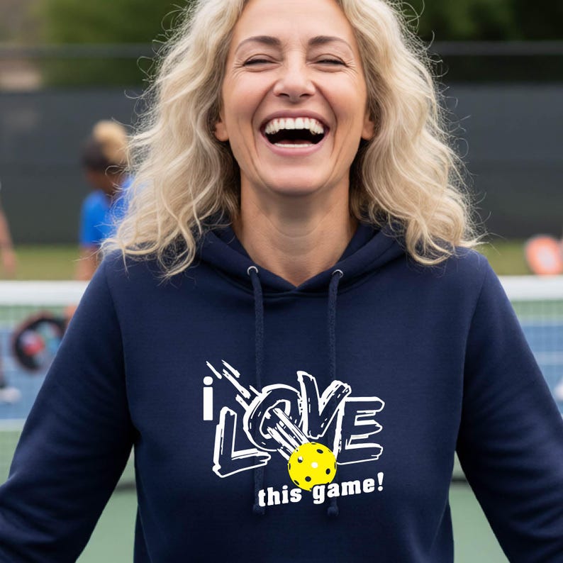 A navy blue hooded sweatshirt featuring a white graphic design that reads "I LOVE this game!" with a yellow pickleball replacing the letter "O". The hoodie includes a matching drawstring cord at the neckline and long sleeves.