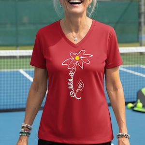 A red V-neck t-shirt featuring a white floral design with the word "Pickleball" written vertically. The flower has a yellow center. The shirt is short-sleeved.
