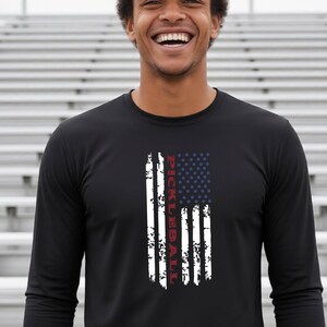 A black long-sleeve shirt featuring a vertical, distressed graphic of the American flag in white, red, and blue. The word PICKLEBALL is printed vertically in red capital letters within the white stripes of the flag design.