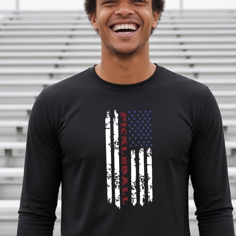 A black long-sleeve shirt featuring a vertical, distressed graphic of the American flag in white, red, and blue. The word PICKLEBALL is printed vertically in red capital letters within the white stripes of the flag design.