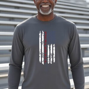 A charcoal grey long-sleeve shirt featuring a distressed graphic of an American flag with vertical white stripes and a blue field. The word PICKLEBALL is printed in red, vertically oriented within the center of the flag design.