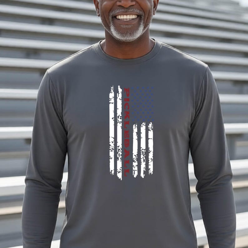 A charcoal grey long-sleeve shirt featuring a distressed graphic of an American flag with vertical white stripes and a blue field. The word PICKLEBALL is printed in red, vertically oriented within the center of the flag design.