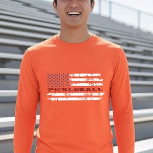 A bright orange long-sleeve shirt featuring a distressed white and blue graphic of the American flag. The word PICKLEBALL is printed in a bold, dark font across the center of the flag design, which has a weathered, textured appearance.