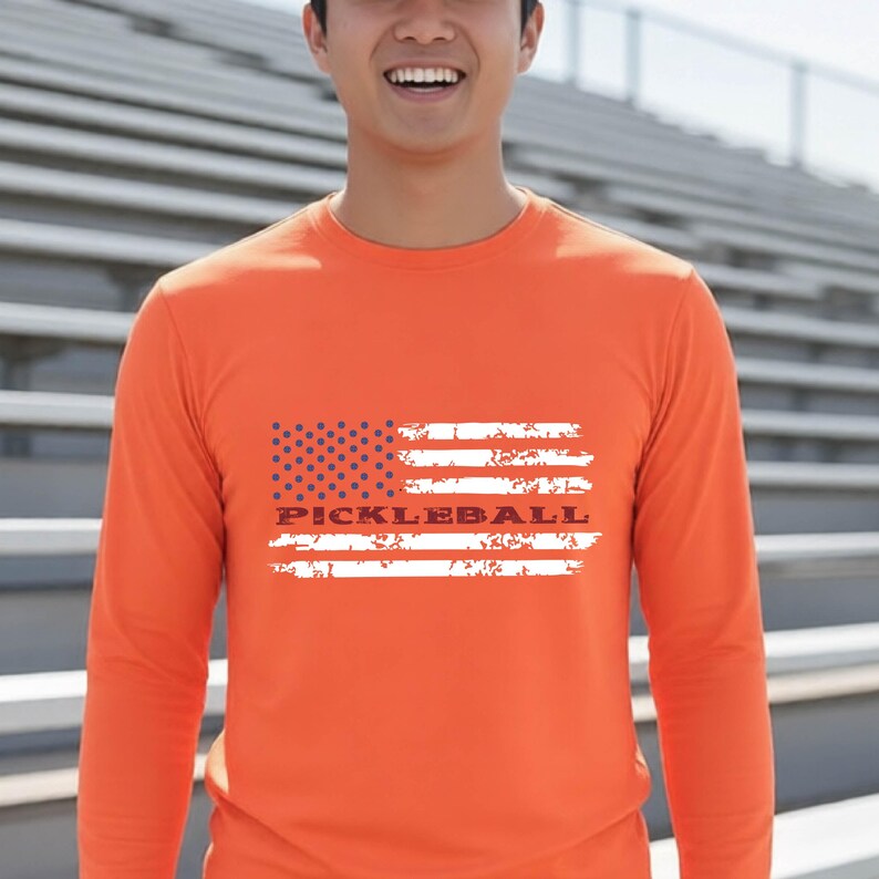A bright orange long-sleeve shirt featuring a distressed white and blue graphic of the American flag. The word PICKLEBALL is printed in a bold, dark font across the center of the flag design, which has a weathered, textured appearance.