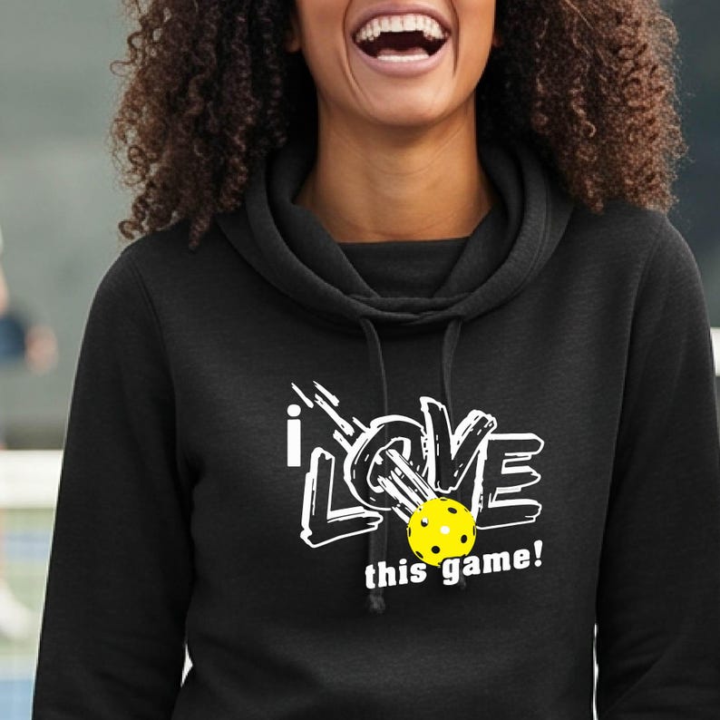A black hooded sweatshirt featuring a white graphic design that reads I LOVE this game! with a yellow pickleball replacing the letter O. The hoodie has a cowl neckline and black drawstrings hanging from the collar area.