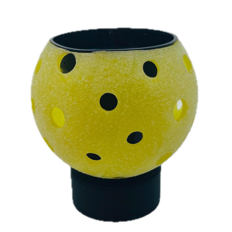 May include: A spherical yellow pickleball with a textured surface and multiple circular black holes, mounted on a solid black cylindrical base. The object features a bright yellow exterior with a series of evenly spaced perforations.
