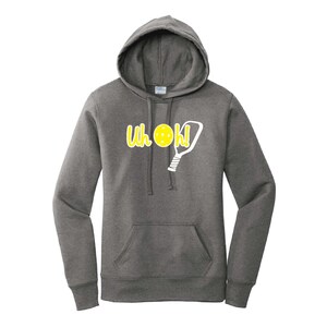 Op de afbeelding: Een donkergrijze hoodie met een voorzak en een trekkoord. De hoodie heeft de tekst "Uh Oh!" in het geel, samen met een gele pickleball en een witte pickleball paddle graphic. De hoodie is gemaakt van een zacht materiaal.