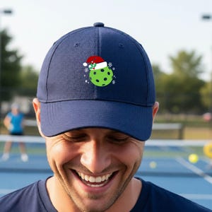 Könnte beinhalten: Eine marineblaue Baseballkappe mit einer Grafik eines hellgrünen Pickleballs, der eine rot-weiße Weihnachtsmütze mit einem Stechpalmenzweig trägt. Das Design ist auf der Vorderseite der Kappe von kleinen weißen Schneeflocken und Sternen umgeben.