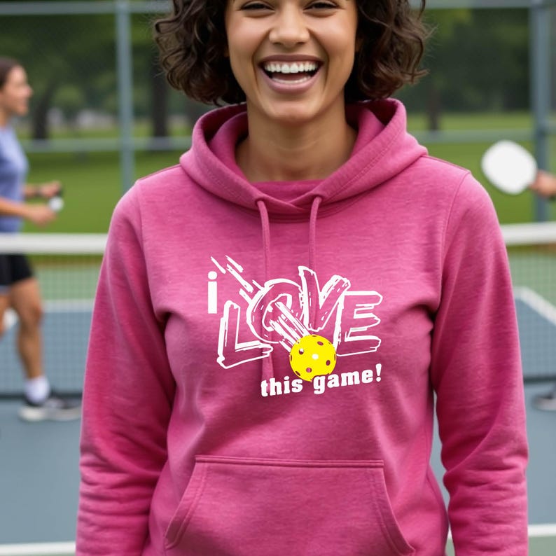 A pink hooded sweatshirt featuring a white graphic design that reads "I LOVE this game!" with a yellow pickleball replacing the letter "O". The hoodie includes a matching drawstring cord at the neckline and long sleeves.
