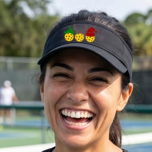 Könnte beinhalten: Ein schwarzer Sonnenschutz mit einer Grafik von drei gelben Pickleballs, die horizontal angeordnet sind. Der erste Ball trägt eine grüne Elfenmütze, der zweite ein braunes Rentiergeweih und der dritte eine rote Weihnachtsmannmütze.