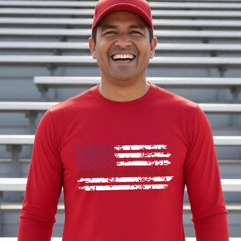 A red long-sleeve shirt featuring a distressed white American flag graphic on the chest with the word PICKLEBALL printed in dark letters across the center of the stripes. The shirt is paired with a matching red baseball cap.