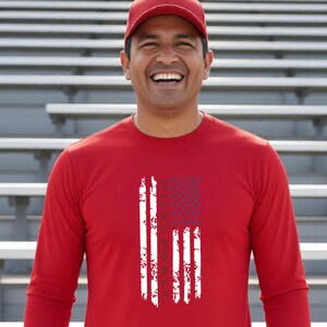 A long-sleeve red shirt featuring a vertical graphic design that resembles a distressed American flag with the word PICKLEBALL integrated into the stripes. The shirt is paired with a matching red baseball cap worn on the head.