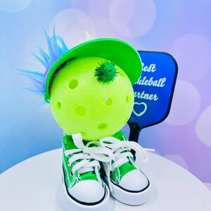May include: A green pickleball with a green hat and a blue paddle that says "Best Pickleball Partner". The pickleball is wearing green sneakers with white laces.