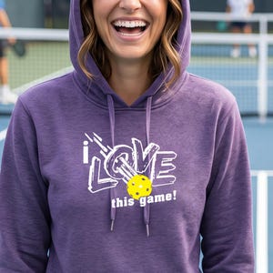 A heathered purple hooded sweatshirt featuring a white graphic that reads I LOVE with a yellow pickleball replacing the letter O, followed by the text this game! in white. The hoodie includes matching purple drawstrings at the neck.