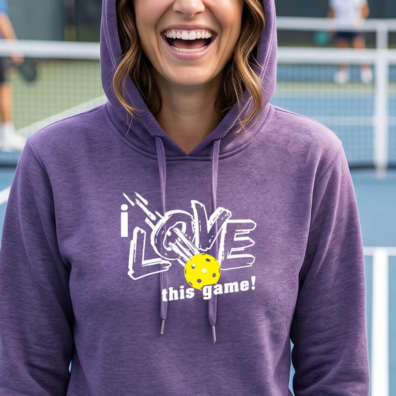 A heathered purple hooded sweatshirt featuring a white graphic that reads I LOVE with a yellow pickleball replacing the letter O, followed by the text this game! in white. The hoodie includes matching purple drawstrings at the neck.
