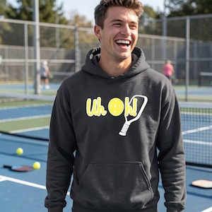 A dark gray hooded sweatshirt featuring a white and yellow graphic on the chest. The graphic displays the text Uh oh! with a yellow pickleball replacing the letter o and a white pickleball paddle outline next to the exclamation point.