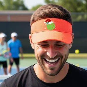 Könnte beinhalten: Ein orangefarbenes Visier mit einer Grafik eines grünen Pickleballs, der eine rot-weiße Weihnachtsmütze mit einem kleinen Zweig Stechpalme trägt. Das Visier hat einen strukturierten Schirm und ein breites Band, das um den Kopf verläuft.