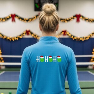 以下が含まれることがあります： ライトブルーの長袖クォータージッププルオーバー。背中には「HOHOHO」という文字がプリントされており、それぞれの「O」の文字が、赤いサンタ帽子をかぶった緑色のピックルボールに置き換えられたデザインになっています。