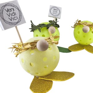 May include: Three yellow, round, decorative gnomes with gold glitter accents. Each gnome has a green wreath around its head and a wooden nose. One gnome holds a sign that says "veni vidi vici".
