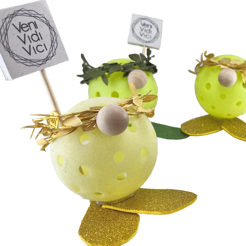 May include: Three yellow, round, decorative gnomes with gold glitter accents. Each gnome has a green wreath around its head and a wooden nose. One gnome holds a sign that says "veni vidi vici".