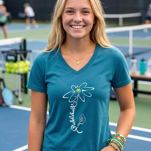 Teal V-neck t-shirt with a white daisy design and the word "Pickleball" in script. The daisy has a rainbow center. A gold necklace with a pickleball paddle pendant is worn.