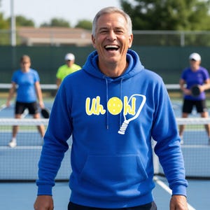 A royal blue hooded sweatshirt featuring a white graphic print on the chest. The design includes the text "Uh oh!" with a yellow pickleball replacing the letter "o" and a white pickleball paddle illustration next to the text.