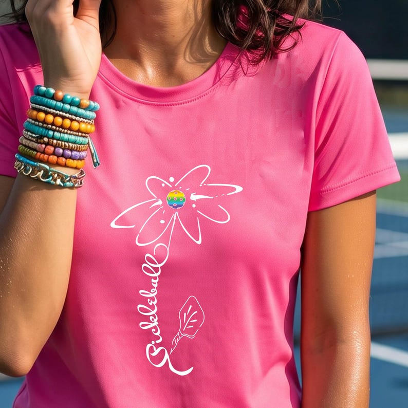 Pink t-shirt featuring a white floral design with a rainbow-colored center and the word "Pickleball" in a script font. The shirt has short sleeves and a crew neck.