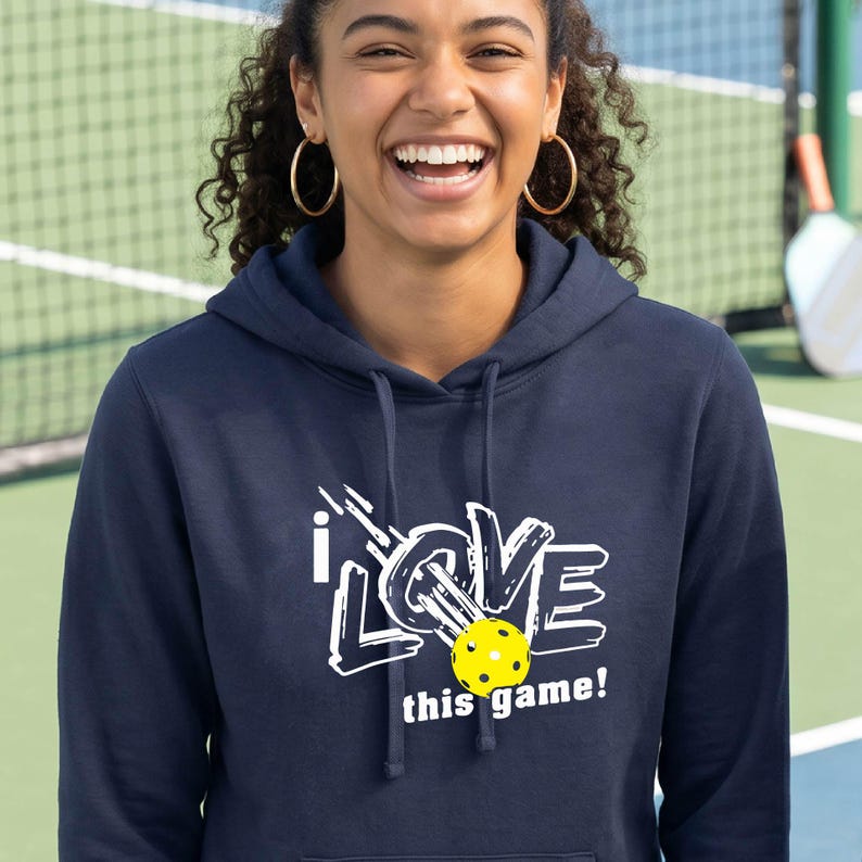 A navy blue hooded sweatshirt featuring a white graphic print that reads I LOVE this game! with a yellow pickleball replacing the letter O. The hoodie includes a drawstring hood and long sleeves, made of a solid dark blue fabric.
