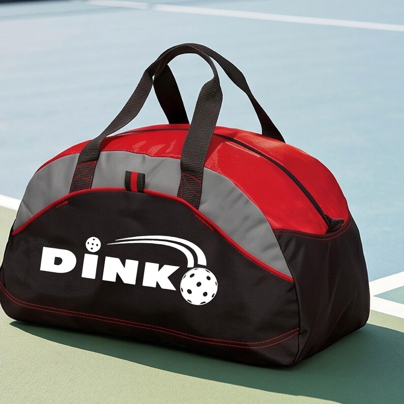 K&ouml;nnte beinhalten: Eine rote, graue und schwarze Sporttasche mit dem wei&szlig;en Schriftzug DINK und einer Pickleball-Grafik. Die Tasche hat schwarze Nylongriffe, ein Hauptfach mit Rei&szlig;verschluss und rote Ziern&auml;hte entlang der Unterkante des schwarzen Bereichs.