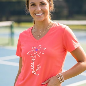 Coral-colored V-neck t-shirt featuring a white flower design with a purple center and the word "Pickleball" in script. The shirt is worn with layered gold necklaces and a beaded bracelet. The background suggests an outdoor setting.