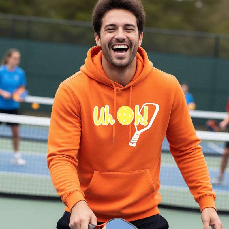 A bright orange hooded sweatshirt featuring a front kangaroo pocket and a graphic print on the chest. The graphic displays the text "Uh oh!" in yellow, accompanied by a white pickleball paddle and a yellow pickleball with holes.