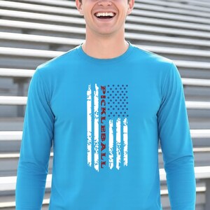 A bright blue long-sleeve shirt featuring a distressed white graphic of an American flag design. The word PICKLEBALL is printed vertically in red capital letters within the center of the flag graphic, replacing the traditional stripes.