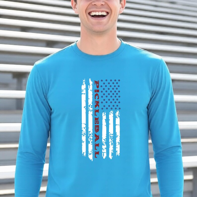 A bright blue long-sleeve shirt featuring a distressed white graphic of an American flag design. The word PICKLEBALL is printed vertically in red capital letters within the center of the flag graphic, replacing the traditional stripes.