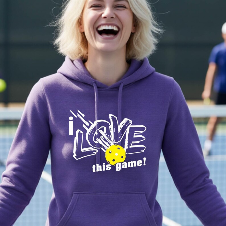 A purple long-sleeve hooded sweatshirt featuring a white graphic design with the text "i LOVE this game!" and a yellow pickleball with black dots replacing the letter O. The hoodie includes a front kangaroo pocket and matching drawstrings.