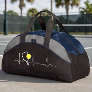 May include: A duffel bag featuring a navy blue top, a grey middle section, and a black base. The front displays a white heartbeat line graphic with a pickleball paddle and a yellow pickleball. It has two black fabric handles for carrying.