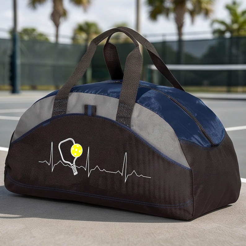 May include: A duffel bag featuring a navy blue top, a grey middle section, and a black base. The front displays a white heartbeat line graphic with a pickleball paddle and a yellow pickleball. It has two black fabric handles for carrying.
