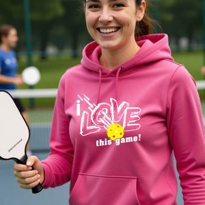 A pink hooded sweatshirt featuring a white graphic design that reads "I LOVE this game!" with a yellow pickleball replacing the letter "O". The hoodie includes a matching drawstring cord at the neckline and long sleeves.