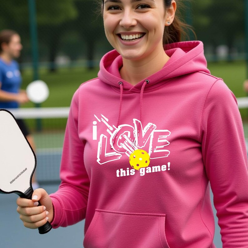 A pink hooded sweatshirt featuring a white graphic design that reads "I LOVE this game!" with a yellow pickleball replacing the letter "O". The hoodie includes a matching drawstring cord at the neckline and long sleeves.