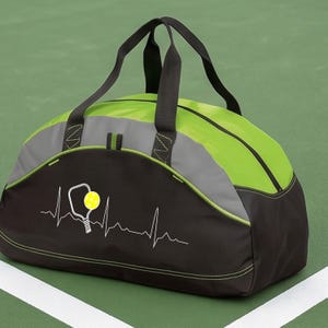 May include: A black and lime green duffel bag featuring a white heartbeat line graphic that incorporates a pickleball paddle and a yellow pickleball. The bag has two black top handles, a zipped main compartment, and grey accent panels.