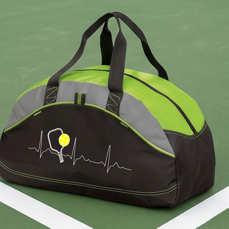 May include: A black and lime green duffel bag featuring a white heartbeat line graphic that incorporates a pickleball paddle and a yellow pickleball. The bag has two black top handles, a zipped main compartment, and grey accent panels.