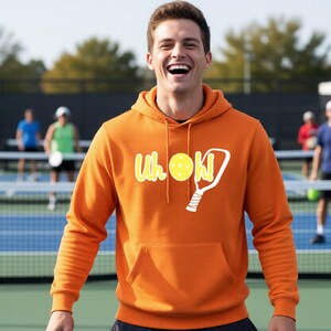 An orange long-sleeve hooded sweatshirt featuring a front kangaroo pocket and a graphic print on the chest. The graphic displays the text "Uh oh!" in white with a yellow pickleball and a white pickleball paddle illustration.