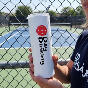May include: A white cylindrical tumbler with a clear lid and a metal straw. The tumbler features a red pickleball graphic above the black text "Day Dinking" printed vertically. The design is centered on the smooth surface of the drinkware.